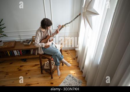 Giovane donna che suona melodia su una chitarra ukulele di legno, seduta davanti alla finestra in un accogliente soggiorno. Foto Stock