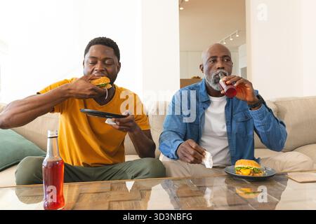 Mangiare hamburger e sorseggiare bibite afroamericane sul divano beige nel soggiorno Foto Stock