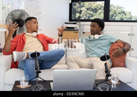 Parlare di diversi amici maschi registrare podcast sul divano in studio domestico, con microfoni e computer portatile Foto Stock
