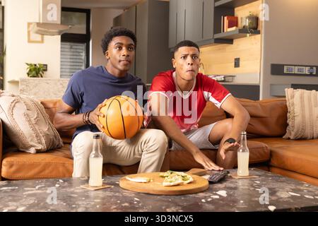 Siediti con diversi amici maschi che si concentrano sulla TV nel soggiorno, con pallacanestro e panini Foto Stock