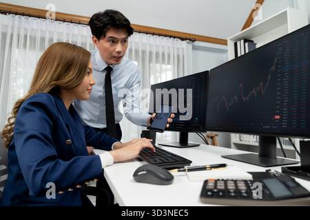 Uomo d'affari asiatico sorridente che mostra dati dinamici del mercato azionario al telefono con una donna che cerca al computer per analizzare il tasso di cambio del valore del profitto Foto Stock