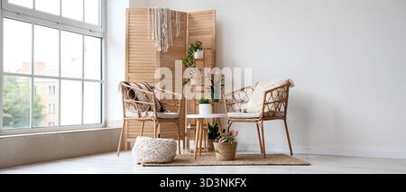 Interno di una stanza luminosa con poltrone in vimini, schermo pieghevole in legno e piante Foto Stock