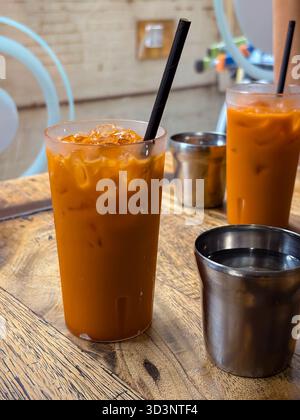 Tè thailandesi ghiacciati, noti anche come Cha yen, in alti bicchieri trasparenti con cannucce nere si siedono su un rustico tavolo in legno accanto a piccole tazze in acciaio inossidabile. Popu Foto Stock