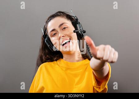 Formazione online. Ritratto di una giovane donna sorridente che indossa le cuffie, con un pollice in su. Sfondo grigio. Il concetto di corsi online e apprendimento Foto Stock