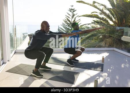 Padre e figlio afroamericani che si esibiscono in squat sul balcone illuminato dal sole, con bottiglie d'acqua Foto Stock