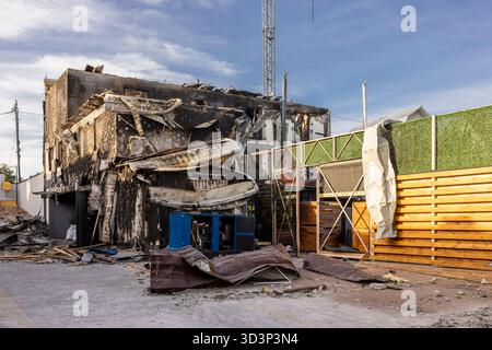 Vista aerea del tetto e delle pareti dell'edificio danneggiati da incendi facciata bruciata crollata e detriti circondano la recinzione intatta del generatore blu e la siepe a contrasto Foto Stock