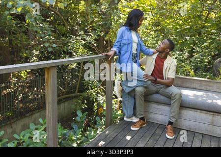 Appoggiandosi su ringhiere, coppia afroamericana che condivide risate su un ponte di legno con panca, copia spazio Foto Stock