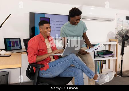 Ridendo colleghi di sesso maschile diversi che scrivono su un notebook in un ufficio moderno, rivedendo i grafici a colori stampati Foto Stock