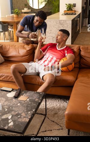 Bere bevande in bottiglia, diversi amici maschi seduti sul divano nel soggiorno, con pallacanestro Foto Stock