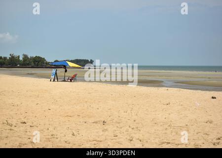 Port Dickson, Malesia - 21 settembre 2025: Una tranquilla spiaggia si dispiega con due persone che si rilassano sotto un grande ombrello blu e giallo. Posi Foto Stock