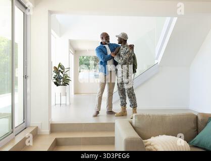 Abbracciare padre e figlio afroamericani che condividono l'abbraccio all'ingresso, con zaino militare e pianta Foto Stock