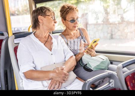 Due amiche mature si siedono insieme su un autobus pubblico, godendosi la compagnia reciproca. Una donna è fidanzata con il suo smartphone mentre l'altra guarda Foto Stock