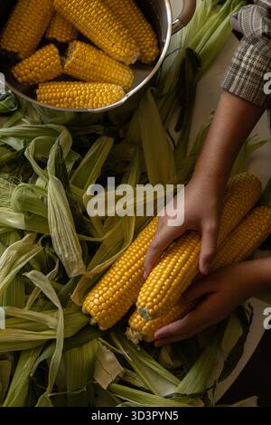 Vista dall'alto di una preadolescente corta e irriconoscibile a casa in cucina, che tiene le pannocchie di mais fresche circondate da bucce durante la preparazione di un pasto. Lo scen Foto Stock
