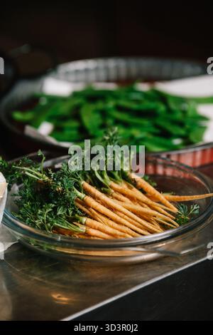Uno shot ravvicinato di carote fresche con cime verdi vivaci in una ciotola rotonda di vetro, collocata su un banco da cucina in acciaio inox, pronta per una sana preparazione Foto Stock