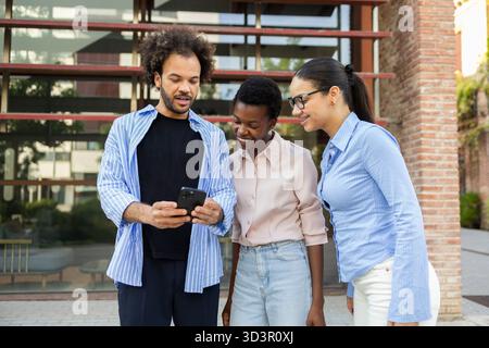 Un team aziendale diversificato esamina il proprio progetto su uno smartphone mentre si trova all'esterno di un moderno edificio urbano. Lo sfondo della città mette in risalto la loro dinam Foto Stock