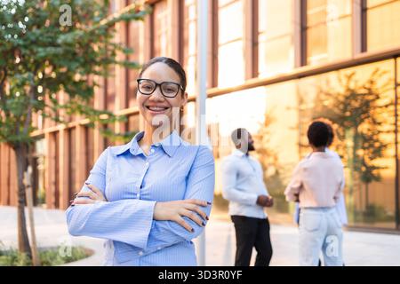 Un team di lavoro sicuro si riunisce all'aperto in un ambiente urbano moderno. L'attenzione è rivolta a un professionista sorridente davanti a sé, con colleghi impegnati Foto Stock