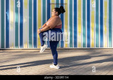 Una donna allunga una gamba e si equilibra sull'altra su uno sfondo vibrante e a righe. Vestita con abbigliamento atletico, incarna fitness e conf Foto Stock