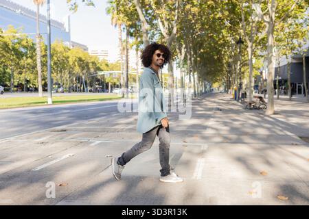 Un uomo con i capelli ricci cammina tranquillamente lungo una strada urbana illuminata dal sole, incarnando uno stile di vita moderno. Vestito con un abbigliamento casual, si diverte a trascorrere una giornata nel V Foto Stock