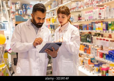 Due farmacisti in camici bianchi esaminano le informazioni su una compressa digitale in una farmacia. Ripiani pieni di vari prodotti farmaceutici sono visi Foto Stock