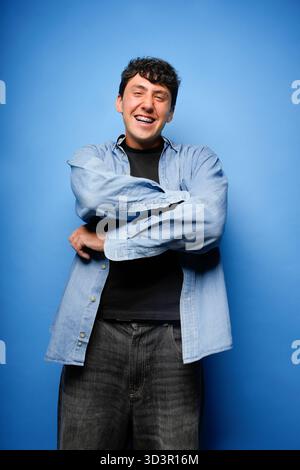 Un giovane gioioso con capelli scuri ricci sorride in grande, davanti a uno sfondo blu brillante. Indossa una camicia blu casual sopra un blac Foto Stock