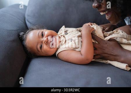 Una madre afroamericana gioiosa e sua figlia condividono un momento giocoso sul divano di casa. Il bambino ride di cuore mentre viene solletico, crea Foto Stock