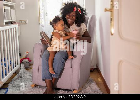 Una madre e una figlia afroamericana condividono un momento di gioia a casa, seduti insieme in una stanza accogliente. La madre tiene un drink mentre la figlia re Foto Stock