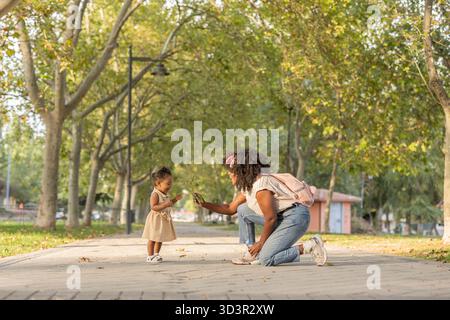 Madre e figlia afroamericane che si godono un momento di gioco in un parco alberato. La madre si inginocchia, presentando un ramoscello, mentre il bambino si impegna eccitatamente Foto Stock