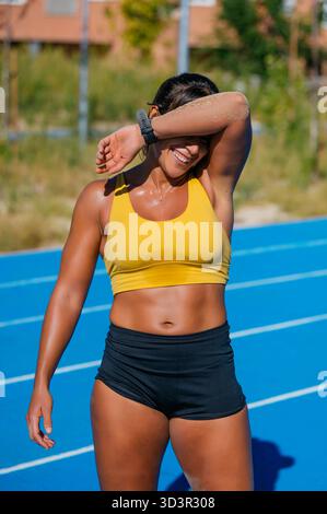 Un'atleta si prende una pausa su una pista all'aperto, appoggiandosi il braccio sulla fronte mentre sorride. Indossa abbigliamento atletico, incarna il fitness e. Foto Stock