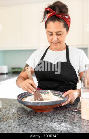 Una donna che indossa un velo rosso e un grembiule mescola l'impasto con un cucchiaio di legno in una ciotola a motivi geometrici, creando pane fatto a mano su un ripiano di granito in un br Foto Stock