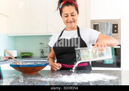 Una donna con grembiule nero e fascia rossa prepara pane fatto a mano su un bancone della cucina. Lei sorride, si concentra sul suo compito, portando un senso di warmt Foto Stock