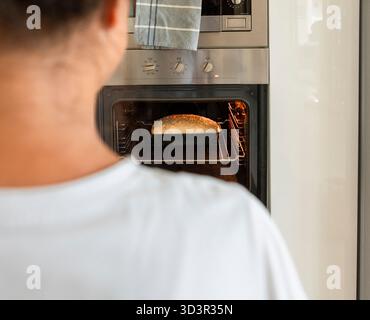 Una donna, vista da dietro, osserva i suoi panini fatti in casa appena preparati nel forno, incarnando il calore e la tradizione della cottura in casa. Foto Stock
