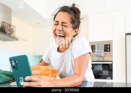 Una donna gioiosa ride di cuore mentre si fa un selfie nella sua cucina moderna, mostrando il suo pane appena sfornato. L'immagine cattura la felicità, Foto Stock