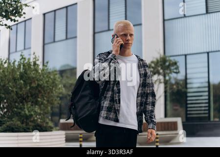 Un giovane dall'aspetto albino, con una camicia a quadri e una maglietta bianca, cammina in un'area urbana mentre parla al telefono, trasmettendo un mese Foto Stock