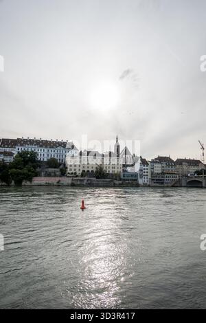 Vista panoramica della città vecchia di Basilea, con edifici storici lungo il fiume sotto un cielo nuvoloso. Il fiume Reno scorre in primo piano, riflettendo il so Foto Stock