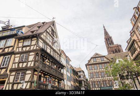 Esplora l'affascinante città vecchia di Strasburgo, in Francia, con le tradizionali case in legno e la maestosa guglia della cattedrale sotto un cielo nuvoloso. Foto Stock