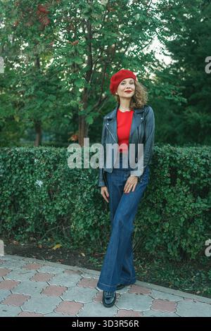 Una donna con un'elegante giacca nera si appoggia su una siepe verde lussureggiante in un vivace parco cittadino. Trasuda eleganza e fiducia, abbracciando la tranquillità Foto Stock