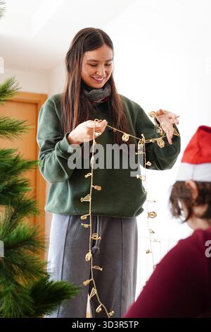 Una ragazza decora con gioia la sua casa per il periodo natalizio, tenendo ghirlande festose accanto a un albero di Natale. Lo spirito natalizio riempie la camera durante il festeggiamento Foto Stock