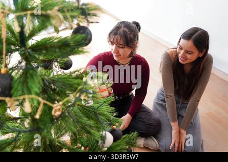 Due donne sedute sul pavimento che decorano con gioia un albero di Natale con ornamenti. L'ambiente trasmette calore e lo spirito festivo del Natale a. Foto Stock