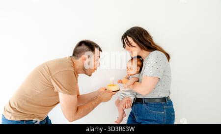Un momento di emozione mentre un padre tiene una piccola torta con candele accese verso una madre sorridente che tiene in braccio il loro adorabile bimbo di due mesi, CA Foto Stock