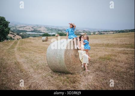 Un momento sereno in un campo di campagna italiano vede una madre e le sue figlie sedute su una balla di fieno. Le colline ondulate e i cieli limpidi creano una pict Foto Stock