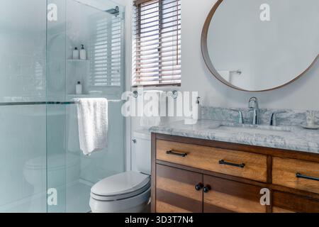 Bagno interno bianco moderno con lavandino in marmo, rubinetto, armadio in legno, doccia in vetro, specchio, e il bagno Foto Stock