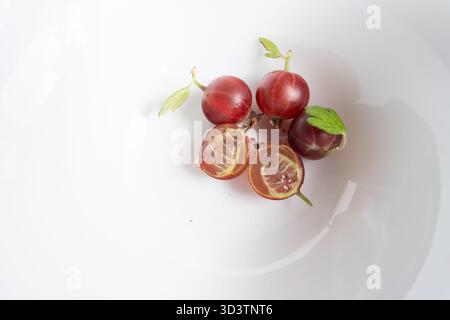 diverse uva spina matura disposte in una ciotola bianca. Una bacca viene tagliata a metà, rivelandone la polpa e i semi. Lo sfondo è una ciotola bianca e pulita Foto Stock