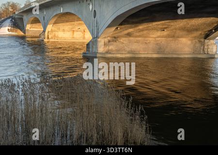 Il fondo del ponte in inverno, le rive innevate, il sole della sera splende sui supporti del ponte, fotografato dall'alto del livello dell'acqua, il ponte i. Foto Stock