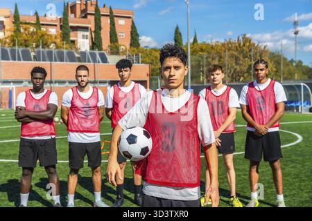 Gruppo eterogeneo di giocatori di calcio maschili in pettorali rossi in piedi su un campo di allenamento, tenendo in mano una palla e pronti per la pratica o per una partita competitiva con concentrazione Foto Stock