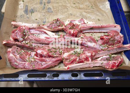 Cibo, costolette di agnello crudo condite con rosmarino ed erbe aromatiche, disposte su un vassoio da forno rivestito in pergamena pronto per la tostatura o la cottura alla griglia Foto Stock