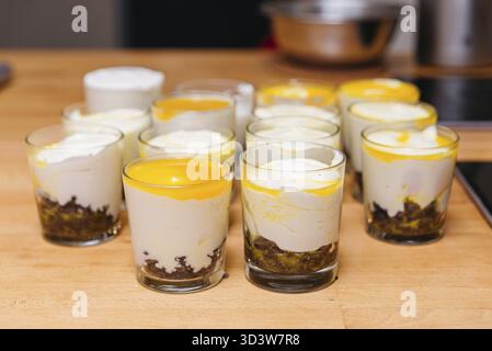 Cibo, Un lotto di dolci cremosi a strati in tazze di vetro trasparente, conditi con purea di mango e sistemati su un banco da cucina in legno Foto Stock