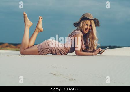 Giovane donna magra in abito e cappello che riposa sulla spiaggia estiva con il telefono in mano Foto Stock
