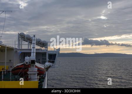 Una nave di pattuglia attraversa il mare calmo al tramonto, mostrando una scialuppa di salvataggio, l'equipaggiamento di soccorso e le strutture del ponte. Nuvole spettacolari diffondono la luce come si forma una terra lontana Foto Stock