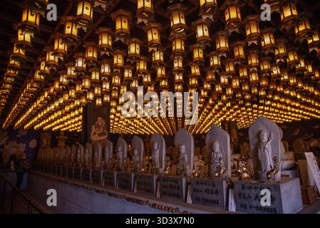 Una sala luminosa piena di centinaia di lanterne appese e file di statue di Buddha all'interno di un tradizionale tempio buddista giapponese. Foto Stock