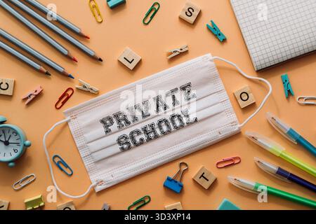 Maschera bianca con la parola Scuola privata scritta sopra. La maschera è posizionata su un tavolo con vari articoli per ufficio, come penne, matite e carta cli Foto Stock